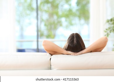 Vista posterior de una mujer relajada sentada en un sofá con las manos en la cabeza y mirando al aire libre a través de la ventana de la sala de estar en casa