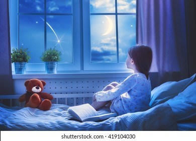Cute child girl sitting at the window and looking at the stars. Girl making a wish by seeing a shooting star at bedtime night.