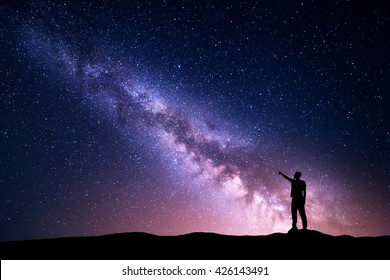 山の夜の星空に立っている若い男の人差し指のシルエットと天の川。夜の風景。宇宙、星と黄色の光でいっぱいの紫の空と旅行の背景