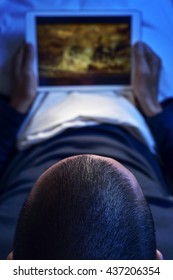 een jonge blanke man die in bed ligt, kijkt 's nachts naar een film of een serie in streaming op zijn tabletcomputer