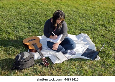 Eine junge Sängerin oder Songwriterin mit ihrer Gitarre und ihrem Computer im Freien im Gras.