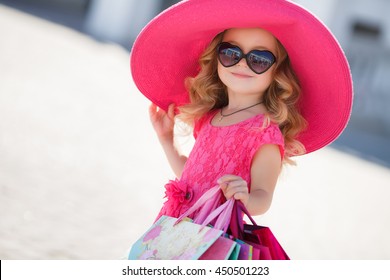 Niña linda con sombrero de moda en sus compras. Retrato de un niño con bolsas de compras. Niño vestido, gafas de sol y zapatos cerca del centro comercial divirtiéndose. compras. niña. Moda