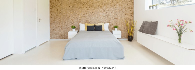 Spacious and bright bedroom interior with the marital bed with cushions, small commodes and wooden wall on a bedhead