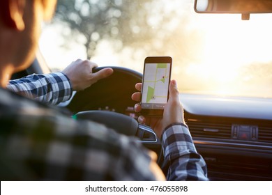 Hombre con camisa a cuadros sentado en el auto y sosteniendo un teléfono móvil negro con navegación gps de mapa, tonificado al atardecer.