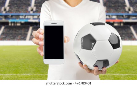 Cropped image of a young football player holding a soccer ball in one hand and the smartphone in another wearing white tshirt at the stadium in Madrid