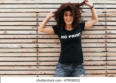 Chica hipster con cabello rizado con camiseta y jeans posando contra la pared de madera, estilo callejero swag