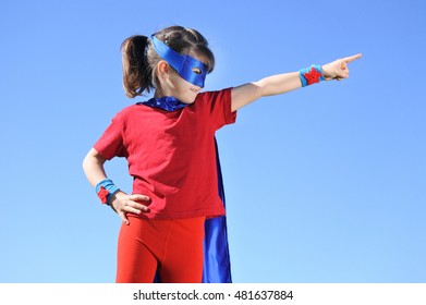 Niña superhéroe (niña de 6 años) señala hacia un fondo de cielo azul dramático con espacio para copiar. Foto conceptual de superhéroe, poder femenino, juego de simulación, infancia, imaginación. Personas reales