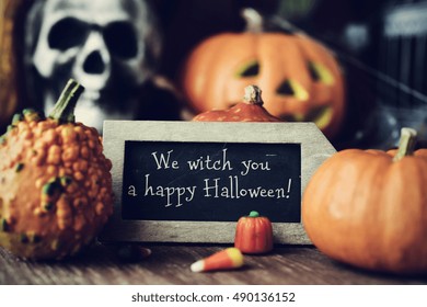 una pizarra con el texto Te esperamos un feliz Halloween, rodeado de diferentes calabazas, colocadas sobre una superficie de madera rústica, y algunos adornos de miedo, como una calavera o una calabaza tallada