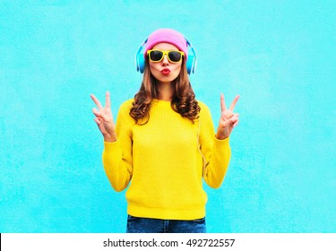 Fashion pretty cool girl in headphones listening to music wearing colorful pink hat yellow sunglasses and sweater over blue background