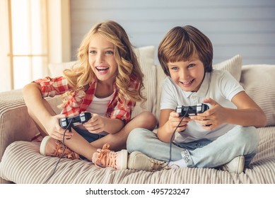 Pretty little girl and boy are playing game console and laughing while sitting on sofa at home