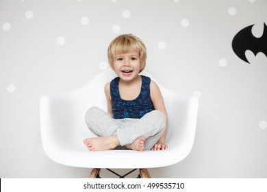 パジャマを着た金髪の遊び好きな幼児のポートレート、あぐらをかいて椅子に座り、カメラ目線、笑い、口を大きく開け、白い歯を見せ、空白の壁にバットマンのロゴが勝つ