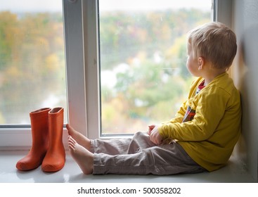Adorable niño rubio sentado en el alféizar de la ventana con zapatos de lluvia y mirando las gotas de lluvia de otoño, en el interior. Un niño mira por la ventana. color de la vendimia botas de goma naranja de pie cerca de la ventana