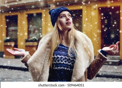 Potret luar ruangan dari wanita muda tersenyum bahagia yang cantik berpose di jalan. Model melihat ke atas, ke langit. Wanita mengenakan pakaian musim dingin yang bergaya. Efek hujan salju ajaib. Natal, konsep tahun baru. pinggang ke atas