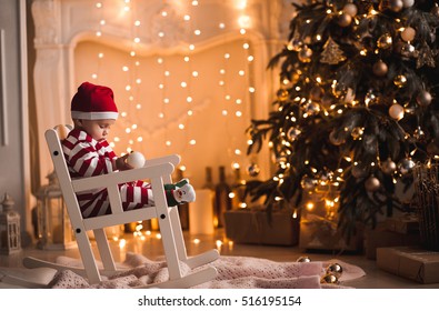 Em bé 1 tuổi mặc bộ đồ ông già Noel ngồi trên ghế bập bênh với cây thông Noel và đèn nền trong phòng. Giáng sinh vui vẻ. Kỳ nghỉ lễ.