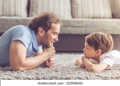 Vista lateral del guapo padre y su lindo hijo mirándose y sonriendo mientras pasan tiempo juntos en casa
