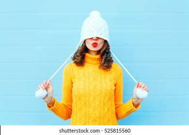 Glückliches kühles Mädchen, das rote Lippen bläst, macht Luftkuss, der eine Strickmütze, gelben Pullover über blauem Hintergrund trägt