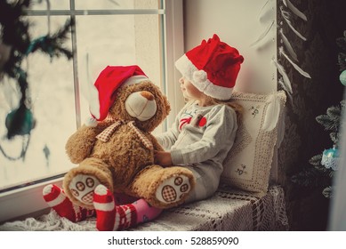 little girl in a Santa Claus hat with teddy bear sitting on the window. The child looks out the window and waiting for Christmas, Santa Claus