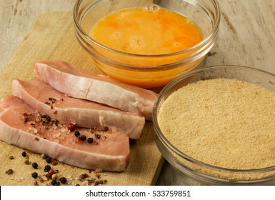 Drei Stücke frische rohe Schweinekoteletts, Schalen mit Semmelbröseln und Eiern zum Panieren des Fleisches, Pfefferkörnern und Salzkristallen. Nahe, flache, horizontale Ansicht von oben