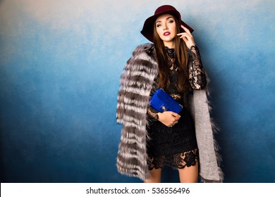 Modny portret atrakcyjnej brunetki, młodej, czarującej kobiety na odizolowanym niebieskim tle. Luksusowa modelka ma modny, puszysty płaszcz i piękny bordowy kapelusz. Moda, mała czarna, celebrytka.