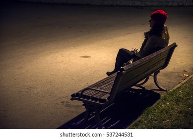 Sad unhappy woman sitting on bench in loneliness and looking away.Stressed woman in a night time.Young sad thoughtful woman sitting on bench against street lamp at night on bokeh copy space background