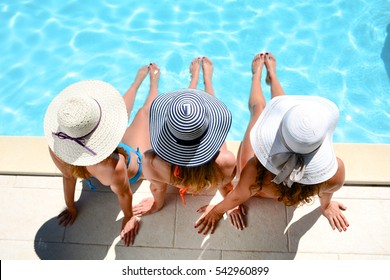 夏の休暇中にリゾートのプールのプールサイドに座っている太陽の帽子を持つ 3 つの美しい若い女性