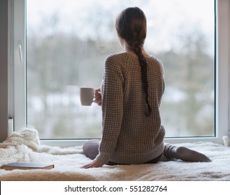 Thoughtful young brunette woman with book and cup of coffee looking through the window, blurry winter forrest landscape outside