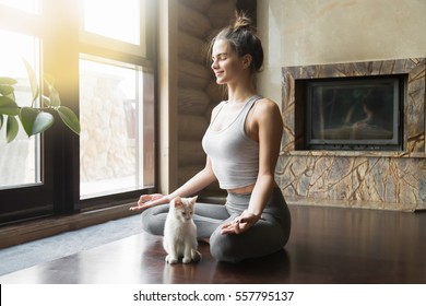 ヨガの練習をしている若い魅力的な笑顔の女性、半蓮の練習、アルダ・パドマサナのポーズ、ワークアウト、スポーツウェアの着用、瞑想セッション、屋内全長、家の内部、近くの猫