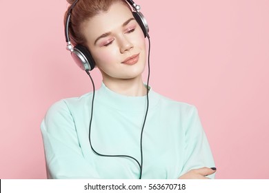 Portrait of a beautiful girl in headphones listening to music smiling with closed eyes standing on a pink background in a blue sweatshirt