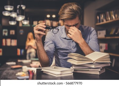 Người thanh niên mệt mỏi vừa cầm kính vừa xoa bóp sống mũi trong thư viện hiện đại đang chăm chỉ làm việc