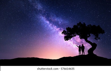 Vía Láctea con gente bajo un árbol en la colina. Paisaje con cielo estrellado nocturno y silueta de un hombre y una mujer felices de pie que señalan con el dedo en el cielo estrellado. Vía Láctea con viajeros. Universo