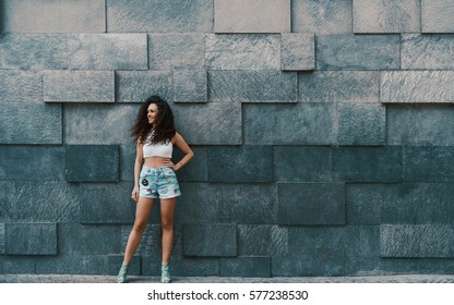 ティール ジーンズのショート パンツで魅力的なカーリー ブルネット ヒップスターの笑顔の女の子通りの前に立って脇を見て、メッセージ、ロゴ、またはプロモーション用のコピー スペースを持つパターン化された壁の背景を投石