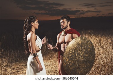 Retrato de mujer morena vestida como grecia con vestido blanco dando amuleto al hombre con pan, guerrera vestida como espartana. Reunión de pareja después de la guerra por la noche.