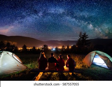 Silhouette of four people sitting on a bench made of logs and watching fire together beside camp and tents in the night. On the background starry sky, Milky way, mountains and luminous town. Rear view