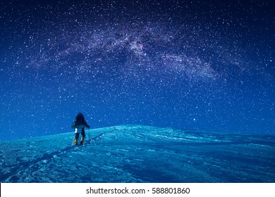 Ein Bergsteiger klettert nachts einen verschneiten Hang hinauf. Milchstraße in einem Sternenhimmel über dem Berggipfel.