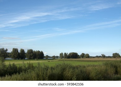 Grüne Landschaft mit Kanal und Baum, Büschen und Feld mit Mais und Gras. Blauer Himmel und weiße Kondensstreifen. Bild von Sonja Riedijk.