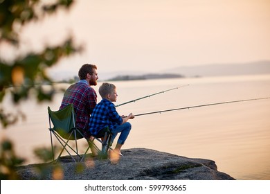 岩の上に一緒に座っている父と息子の横から見たポートレートで、静かな湖の水でロッドを使って釣りをし、夕日の風景があり、両方とも市松模様のシャツを着て、木の後ろから撮影