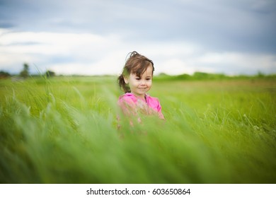 Schattig 3 jaar oud klein brunnete meisje gekleed in roze kleding staat in lang vers groen gras. Baby geniet van de schoonheid van de natuur op het platteland op een bewolkte zomerdag. Horizontale kleurenfotografie.