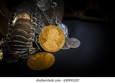 Still Life with two Austria-Hungary thalers, avers and revers of golden coin-ducats from 1915 with Kaiser Franz Joseph I, leaning on silver jewelery and dark environment