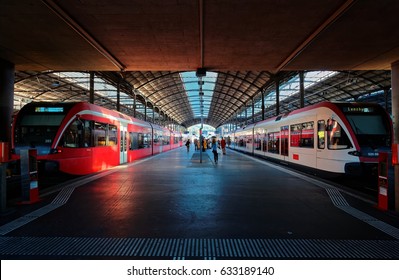 Vista en perspectiva de un andén en la estación central de trenes de Lucerna con la luz del sol proyectada sobre los trenes que se estacionan junto al andén y los pasajeros se apresuran a abordar ~ Un hermoso rincón en la terminal de trenes de Lucerna