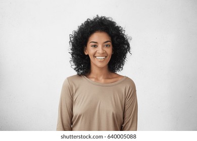 幸せな笑顔でスタジオでポーズをとって巻き毛を持つ陽気な若い混血女性のウエスト アップの肖像画。カジュアルな服装をした浅黒い肌の女性が嬉しそうに微笑み、白いまっすぐな歯を見せている