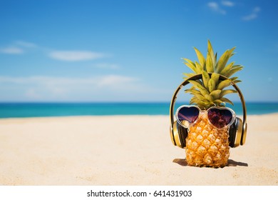 Ripe attractive pineapple in stylish sunglasses heart shape and gold headphones on sand against turquoise  sea water. Tropical summer vacation concept. Summer sunny day on the beach of tropical island