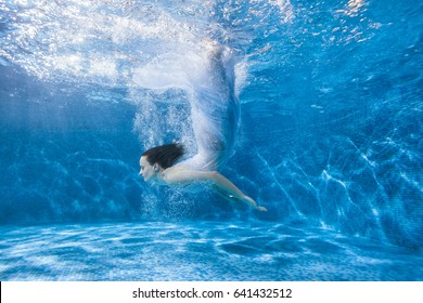 女人美人鱼潜入泳池水下，她身穿白色连衣裙，水花四溅。