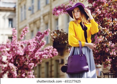 Outdoor portrait of young beautiful girl posing in street. Model wearing stylish hat, shirt, skirt, holding purple bag, handbag. City lifestyle. Female fashion concept. Copy, empty space for text
