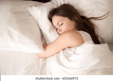 Draufsicht einer attraktiven jungen Frau, die gut im Bett schläft und ein weiches weißes Kissen umarmt. Teenager-Mädchen ruht, gutes Nachtschlafkonzept. Dame genießt frische weiche Bettwäsche und Matratze im Schlafzimmer