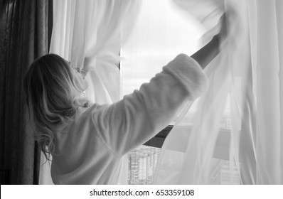 mulher jovem adulta em um manto branco abre as cortinas e fica na janela