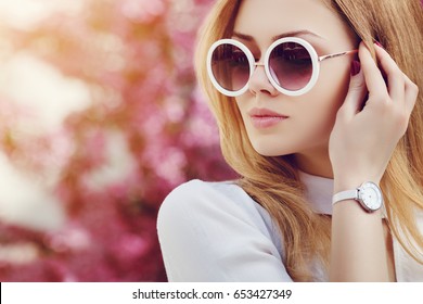 Nahaufnahmeporträt des jungen schönen modischen Mädchens, das in der Straße aufwirft. Modell trägt stilvolle weiße runde Sonnenbrille, Handgelenk, Handuhr. Weibliches Modekonzept.