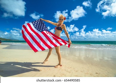 アメリカ、ハワイ、オアフ島の東海岸、壮観な熱帯ラニカイ ビーチでアメリカ国旗を振ってアメリカ国旗ビキニで笑顔の女性。ハワイでの休暇を楽しみ、自由に。愛国的な概念。