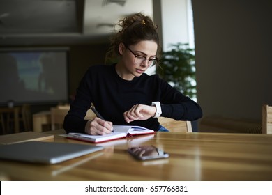 Getalenteerde vrouwelijke schrijver in stijlvolle bril die wat informatie in notitieblok opmerkt terwijl ze op de alarmklok kijkt om tijd te beheren voor de organisatie van het werkproces terwijl ze binnen op de universiteit zit