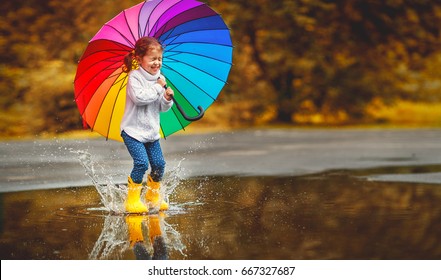 Glückliches lustiges ba Kind durch Mädchen mit einem bunten Regenschirm, der auf Pfützen in Gummistiefeln springt und lacht