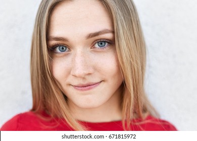 Close up portrait of pretty female with blue alluring eyes, freckled face and pure healthy skin looking directly into camera having thoughtful expression. Cute young female in red clothes posing